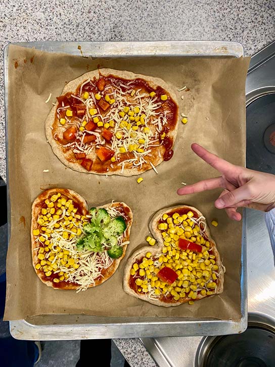 Ein Backblech mit Pizzen in Herzchenform und einer Kinderhand die ein Peace-Zeichen macht